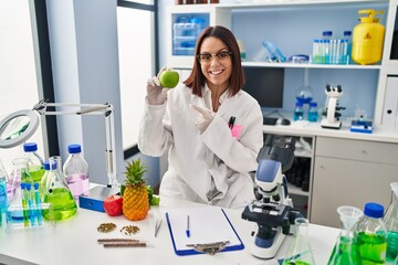 Young hispanic woman working at scientist laboratory holding fruit smiling happy pointing with hand and finger