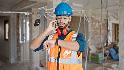 Young hispanic man builder talking on smartphone looking at wrist watch at construction site