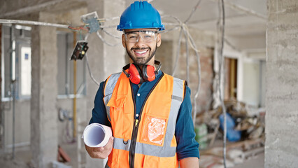 Young hispanic man builder smiling confident standing with blueprints at construction site