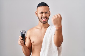 Young hispanic man holding electric razor annoyed and frustrated shouting with anger, yelling crazy with anger and hand raised - obrazy, fototapety, plakaty