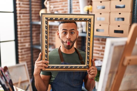 Young hispanic man sitting at art studio with empty frame making fish face with mouth and squinting eyes, crazy and comical.