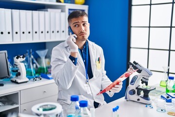 Young hispanic man scientist talking on the smartphone reading document at laboratory