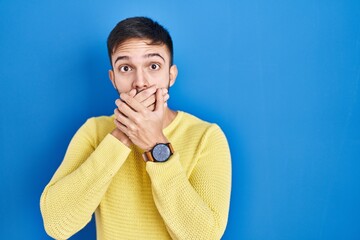 Hispanic man standing over blue background shocked covering mouth with hands for mistake. secret...