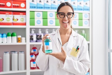 Brunette woman working at pharmacy drugstore holding cough syrup sticking tongue out happy with funny expression.