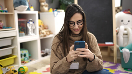 Beautiful hispanic woman working as a teacher using smartphone sitting on floor at kindergarten