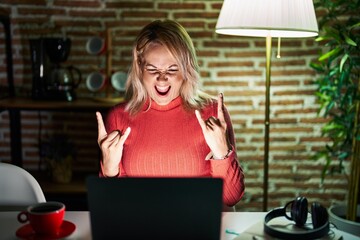 Fototapeta na wymiar Blonde woman using laptop at night at home shouting with crazy expression doing rock symbol with hands up. music star. heavy concept.