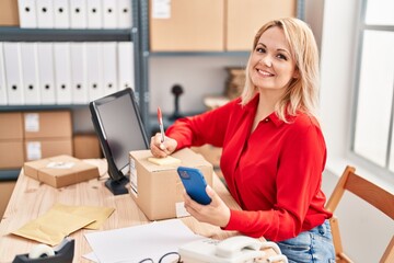 Young blonde woman ecommerce business worker writing on package using smartphone at office