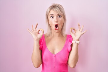 Young caucasian woman standing over pink background looking surprised and shocked doing ok approval symbol with fingers. crazy expression