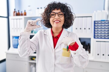 Hispanic doctor woman with curly hair holding vaccine pointing finger to one self smiling happy and proud