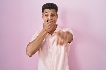 Young hispanic man standing over pink background laughing at you, pointing finger to the camera...