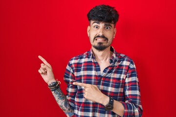 Young hispanic man with beard standing over red background pointing aside worried and nervous with...