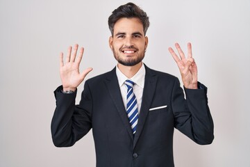 Young hispanic man with tattoos wearing business suit and tie showing and pointing up with fingers number eight while smiling confident and happy.