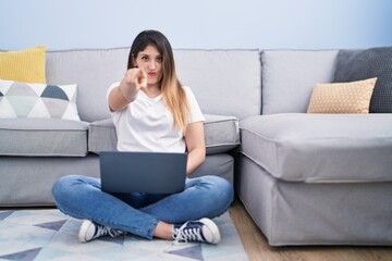 Young brunette woman sitting on the floor at home using laptop pointing with finger to the camera and to you, confident gesture looking serious
