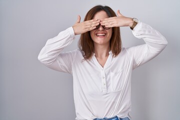 Brunette woman standing over white isolated background covering eyes with hands smiling cheerful and funny. blind concept.