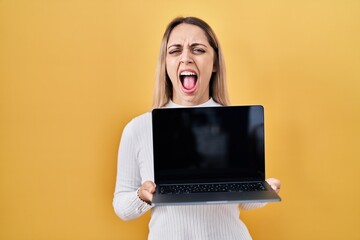 Young blonde woman holding laptop angry and mad screaming frustrated and furious, shouting with...