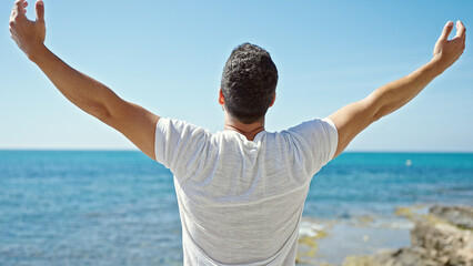 Young hispanic man standing with arms open breathing at the beach