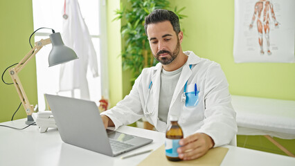 Young hispanic man doctor using laptop holding medication bottle at clinic