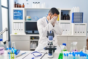 Young hispanic man with beard working at scientist laboratory with sad expression covering face with hands while crying. depression concept.