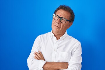 Middle age hispanic man standing over blue background looking to the side with arms crossed convinced and confident