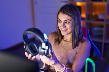 Young beautiful hispanic woman streamer smiling confident holding headphones at gaming room