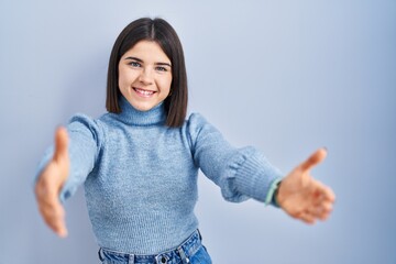 Young hispanic woman standing over blue background looking at the camera smiling with open arms for hug. cheerful expression embracing happiness.