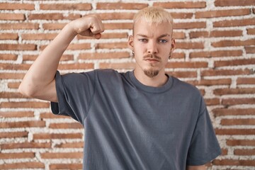 Young caucasian man standing over bricks wall strong person showing arm muscle, confident and proud of power