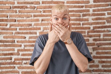 Young caucasian man standing over bricks wall shocked covering mouth with hands for mistake. secret...