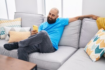 Obraz premium Young bald man using smartphone sitting on sofa at home