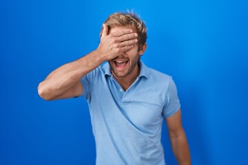 Caucasian man standing over blue background smiling and laughing with hand on face covering eyes for surprise. blind concept.