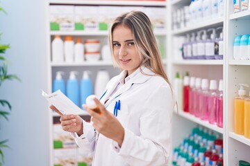 Young blonde woman pharmacist holding pills bottle reading prescription at pharmacy