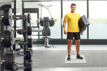 Fototapeta na wymiar Full length portrait of a young man holding an aerobic stepper and smiling at a gym