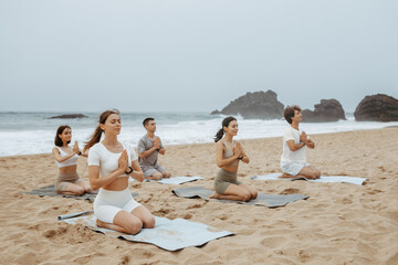 People sitting and meditating on the beach, practicing asana with joined hands, enjoying morning training at coastline