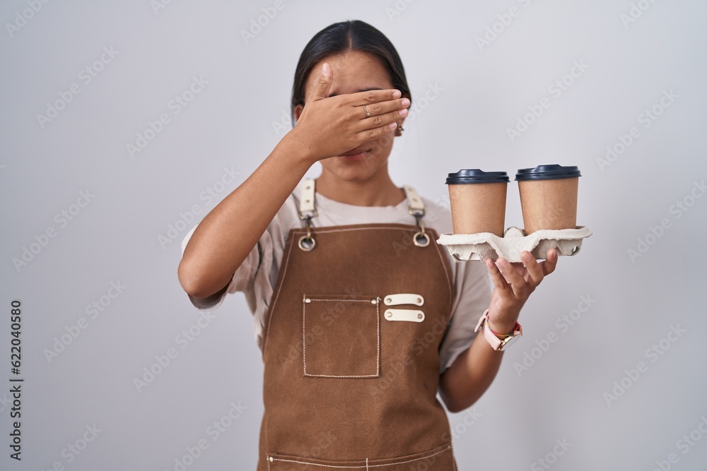 Canvas Prints Young hispanic woman wearing professional waitress apron holding coffee covering eyes with hand, looking serious and sad. sightless, hiding and rejection concept