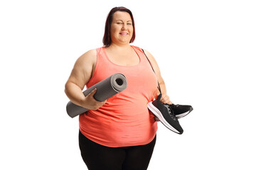 Overweight young woman in sports clothing holding a pair of snickers and an exercise mat