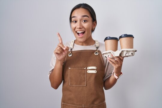 Young hispanic woman wearing professional waitress apron holding coffee pointing finger up with successful idea. exited and happy. number one.