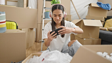 Young beautiful hispanic woman playing video game by smartphone at new home