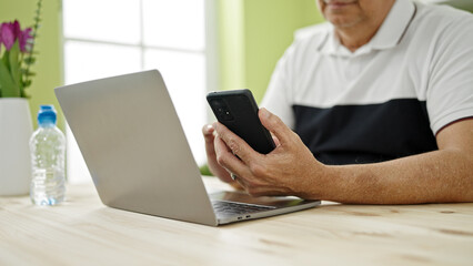 Middle age man with grey hair using smartphone and laptop at dinning room