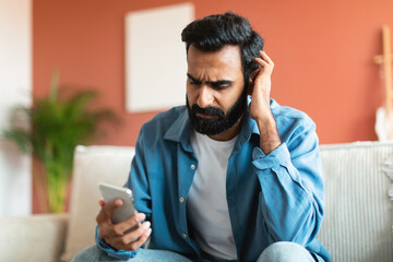 Confused arabic guy looking at smartphone touching head at home