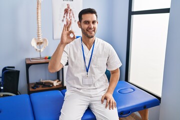 Young hispanic man with beard working at pain recovery clinic smiling positive doing ok sign with hand and fingers. successful expression.