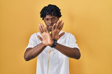 Young african man with dreadlocks standing over yellow background rejection expression crossing...