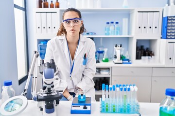 Fototapeta premium Young hispanic woman working at scientist laboratory making fish face with lips, crazy and comical gesture. funny expression.