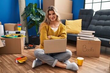 Young woman sitting on the floor at new home using laptop happy face smiling with crossed arms looking at the camera. positive person.