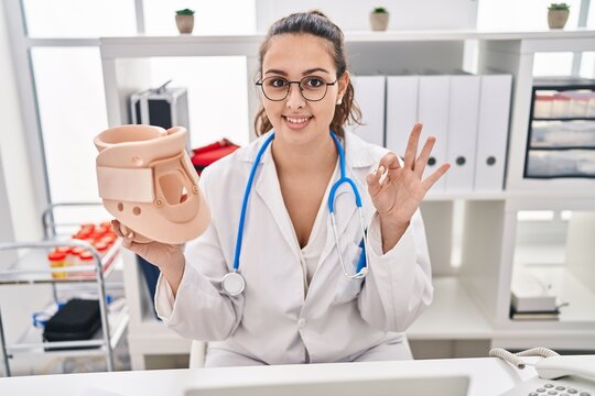 Young hispanic doctor woman holding cervical neck collar doing ok sign with fingers, smiling friendly gesturing excellent symbol