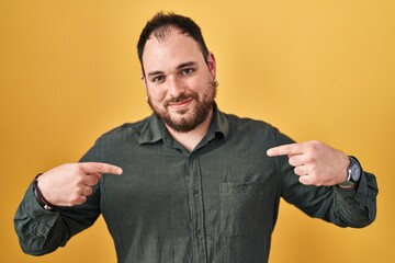 Plus size hispanic man with beard standing over yellow background looking confident with smile on...