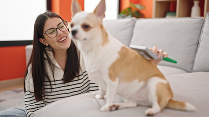 Young hispanic woman with chihuahua dog sitting on the floor brushing dog hair at home