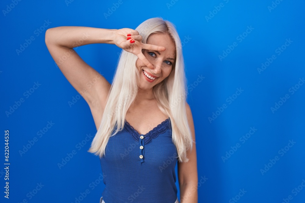 Wall mural Caucasian woman standing over blue background doing peace symbol with fingers over face, smiling cheerful showing victory