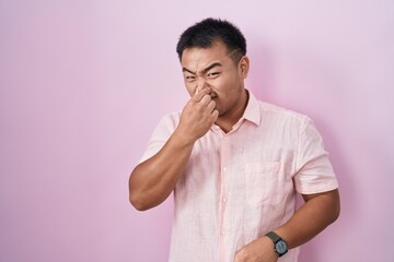 Chinese young man standing over pink background smelling something stinky and disgusting,...