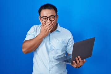 Chinese young man using computer laptop laughing and embarrassed giggle covering mouth with hands,...