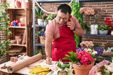 Young chinese man florist talking on smartphone writing on notebook at florist