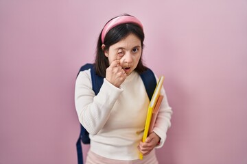 Woman with down syndrome wearing student backpack and holding books pointing to the eye watching you gesture, suspicious expression